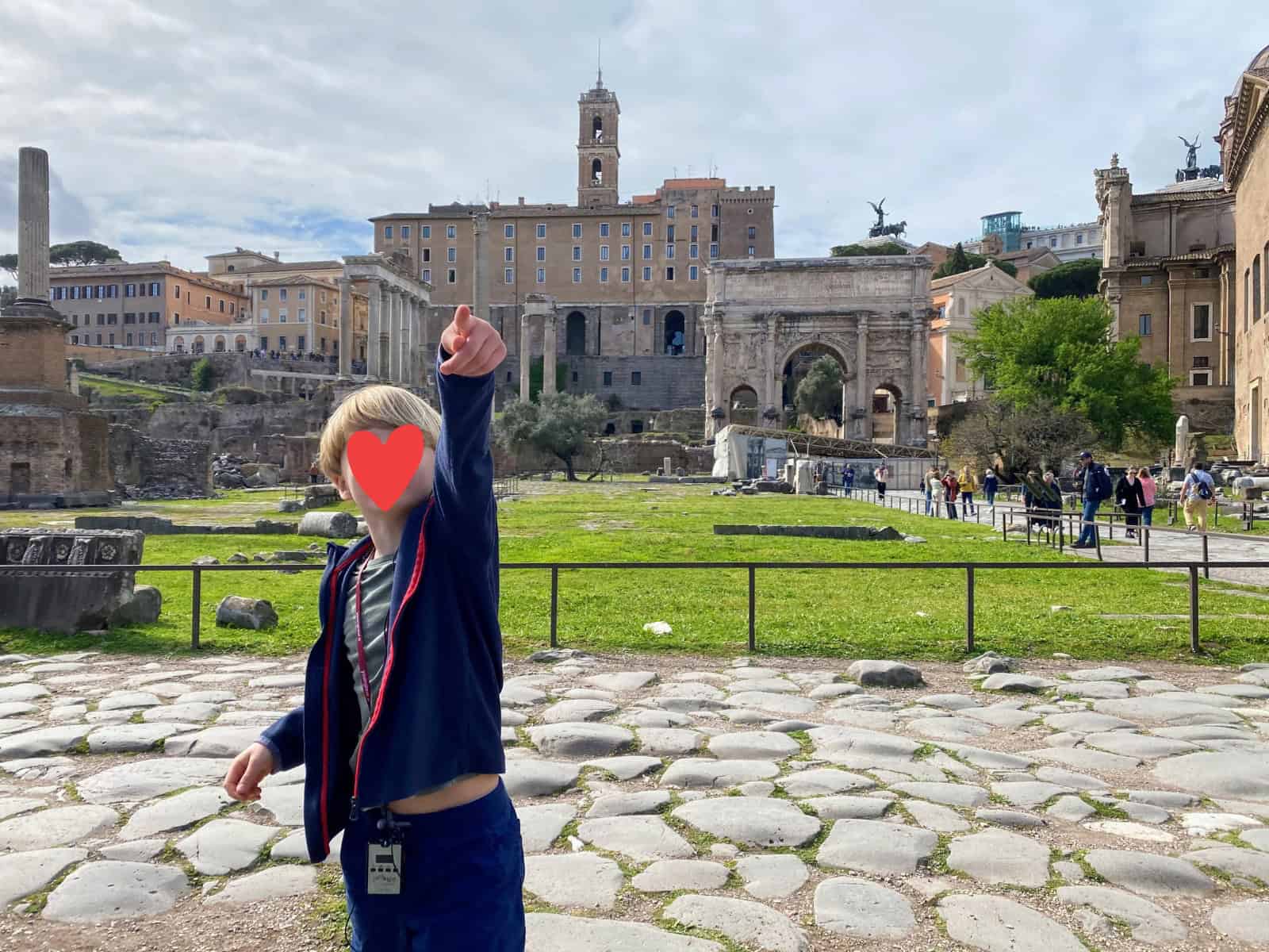 Boy pointing to something at the Roman Forum in Italy. He's standing on cobblestones. Other tourists and parts of the Forum behind him.