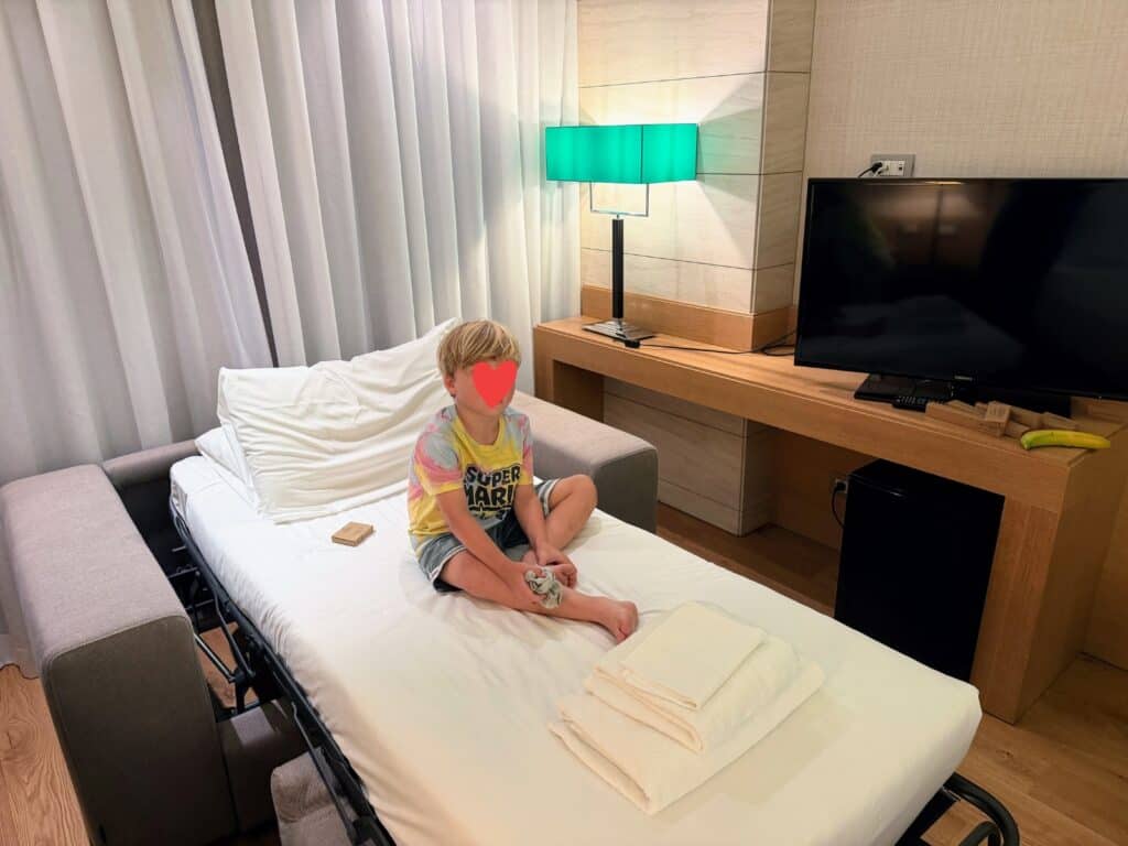 Boy sits on narrow pull out bed at Precise House Mantegna Rome. Lamp with green shade on shelf/desk behind him, along with tv. Banana next to tv. Shades closed behind the bed.