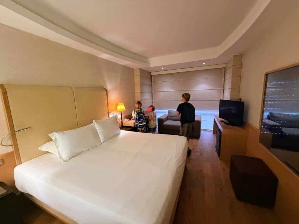 Boys look at table in hotel room with large bed, tv, desk, and lounge chair at Precise House Mantegna Rome. The room has wood, light colors, and the window is covered with a shade.