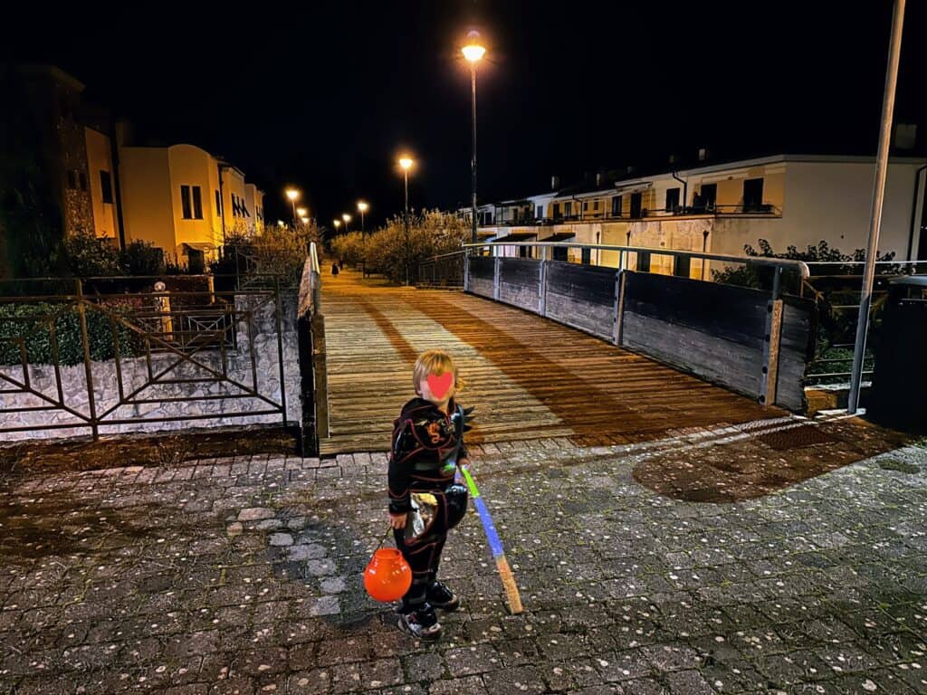 Boy on stone walkway about to cross wooden bridge at night in Italy. He's wearing a ninja costume and carrying a plastic pumpkin bucket and sword.