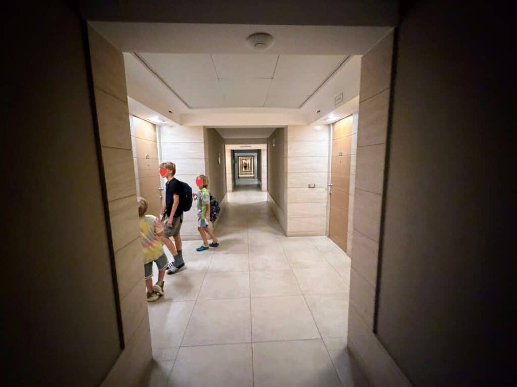 Boys stand in hotel hallway. The hallway is light-colored with stone tile flooring. This is at Precise House Mantegna Rome.