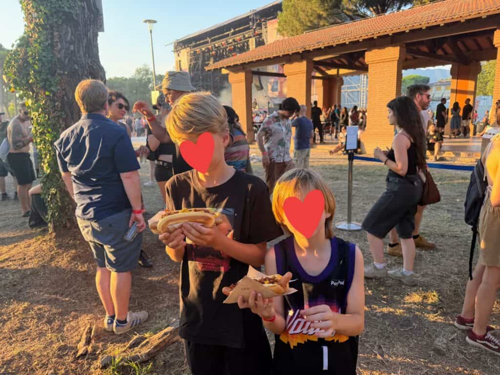 Two boys stand in front of a concert stage in Tuscany, Italy. They are holding hot dogs.