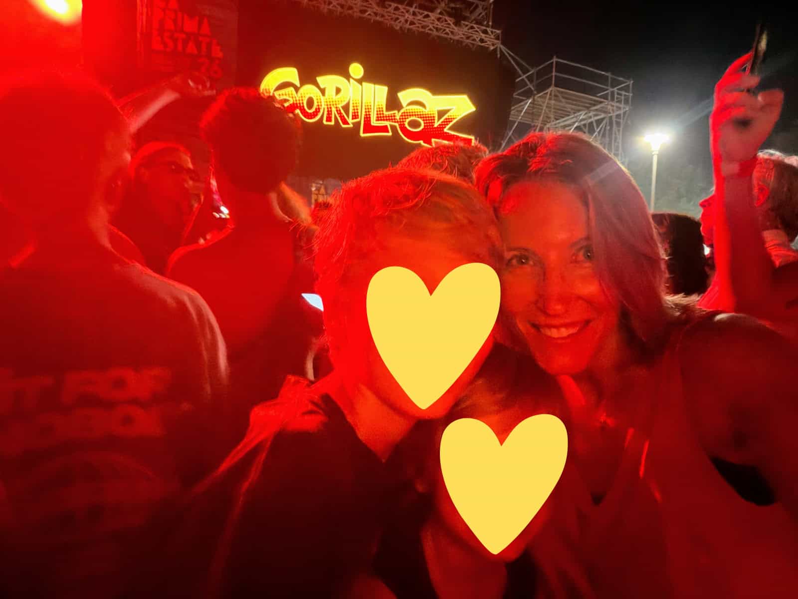 Mom and two sons stand in front of digital sign for 'Gorillaz' at their concert in Tuscany, Italy. They light is red. It's nighttime. Other people stand around them.