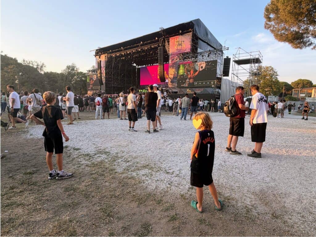 Two boys stand in open dirt/gravel area in front of a large concert stage in Tuscany, Italy. Other people are standing around, most of them closer to the stage. Trees in background.