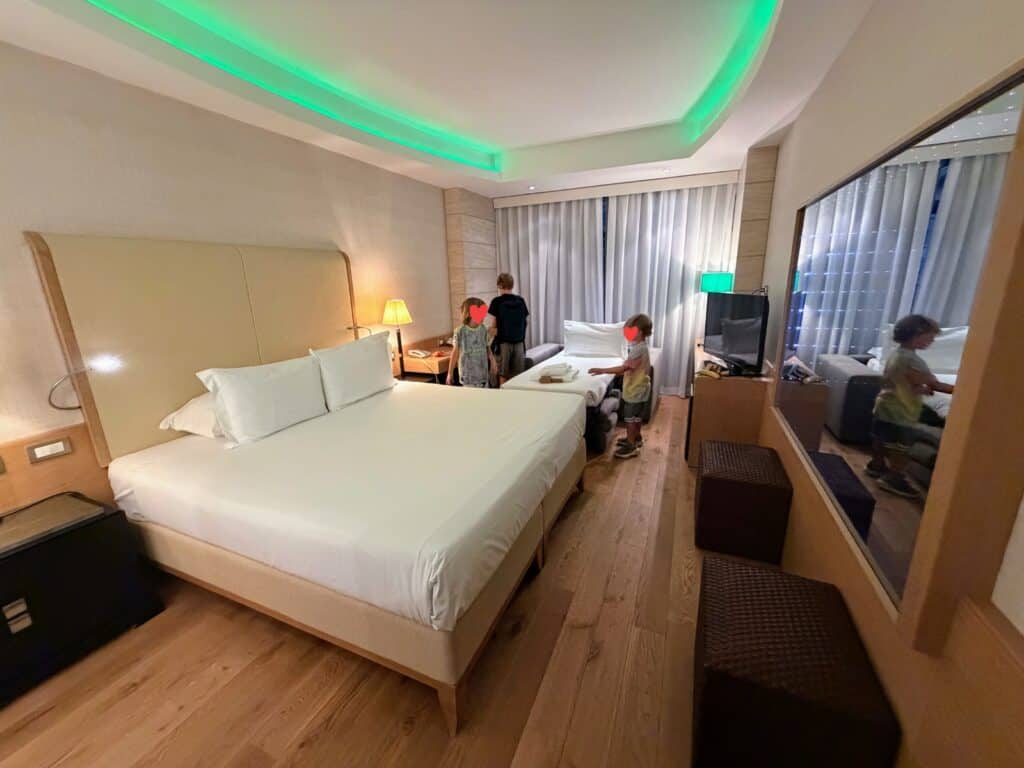 Boys in hotel room at Precise House Mantegna Rome. Green accent lighting on ceiling. Bed in room, behind them the boys look at a small pull out bed and a desk. TV on right and large mirror.