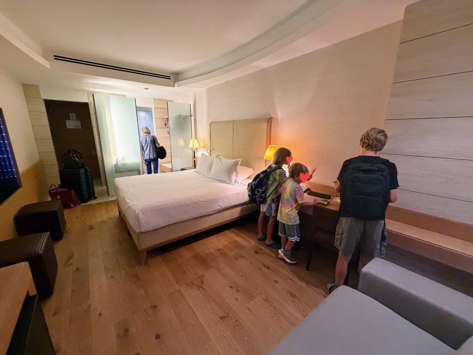 Boys look at desk on right at Precise House Mantegna Rome. Woman walks into bathroom in background. Between woman and boys is a large bed. They are in a light colored, modern hotel room.