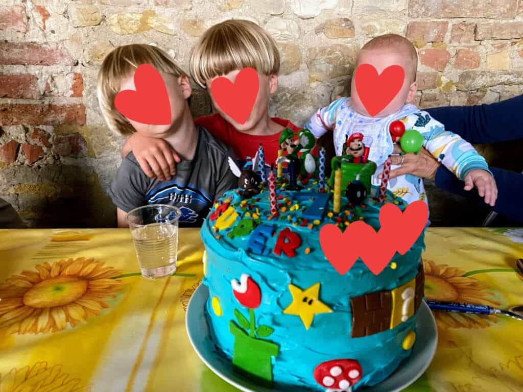 Three young boys sit at the table. There is a Super Mario birthday cake in front of them. Wall behind them is brick.