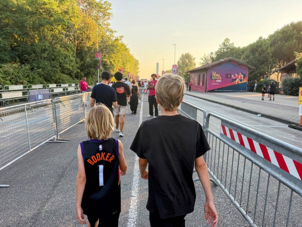 Two boys walking to the entrance to an outdoor concert venue in Tuscany, Italy. Walk is lined with metal fencing on a street cleared of traffic. Trees line both sides of wide street. Pink building with venue advertising ahead on right.