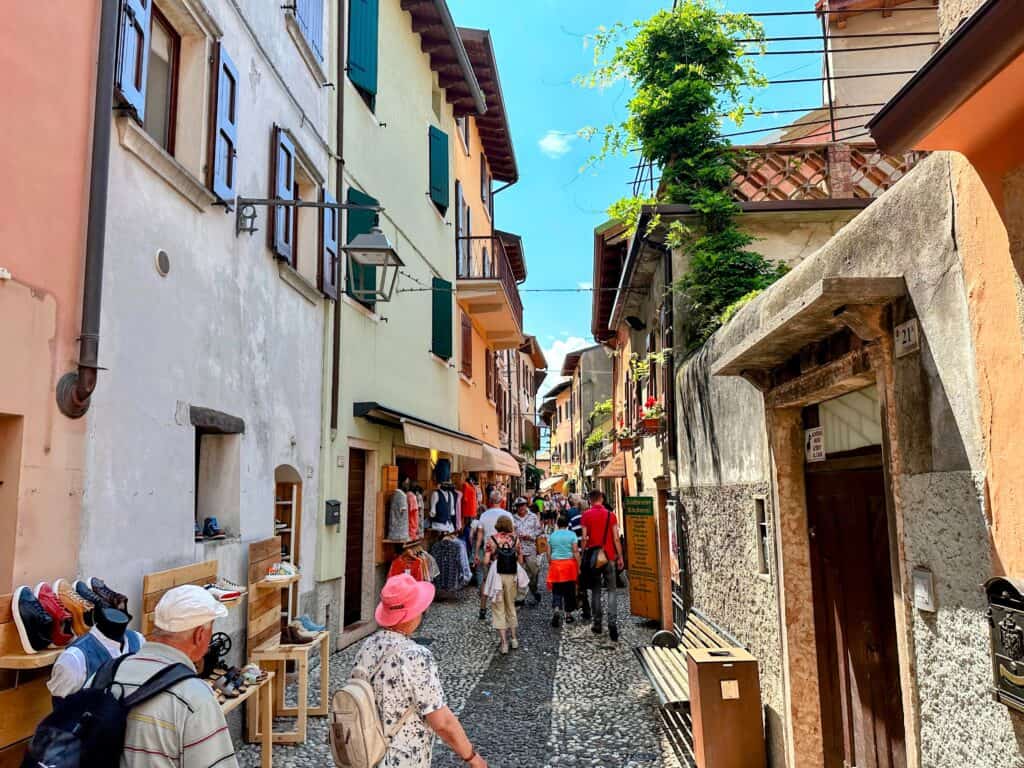 Tourists walk down cobbled lane of Malcesine in Lake Garda. The buildings on either side are painted in colorful pastels. Tourist shops sell shoes and clothes.