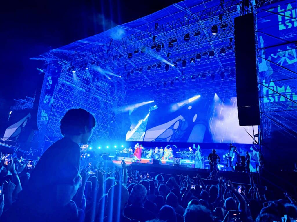 Boy sits on adults shoulders in a crowd of fans in front of a concert stage in Tuscany, Italy. The band The Gorillaz plays on stage.