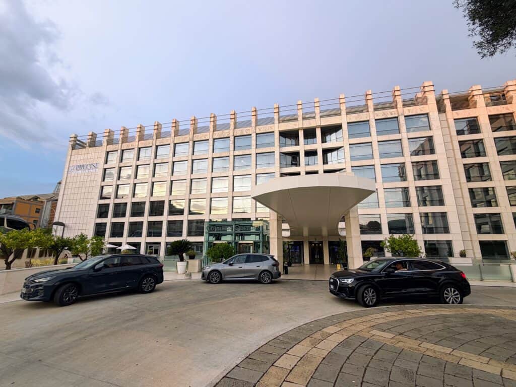 Main entrance to Precise House Mantegna Rome hotel. Three cars parked out front. It's a 6-story building with glass and light colored stone.