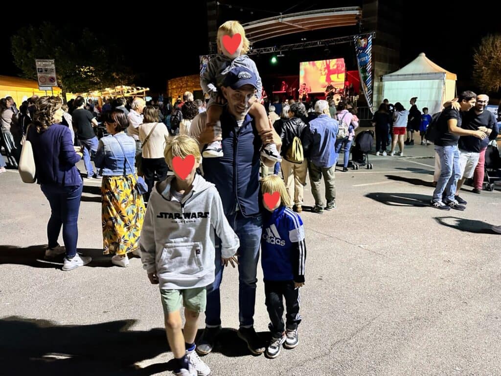 Father and three sons stand in front of concert stage in Tuscany, Italy. Others are closer to the stage. One of the sons is on the father's shoulders.