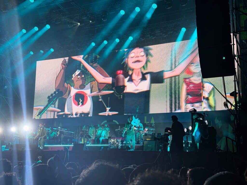 Large screen behind concert stage shows cartoon characters for the band, the Gorillaz. The band is on the stage, playing a song. Fans crowd around the front of the stage below them.