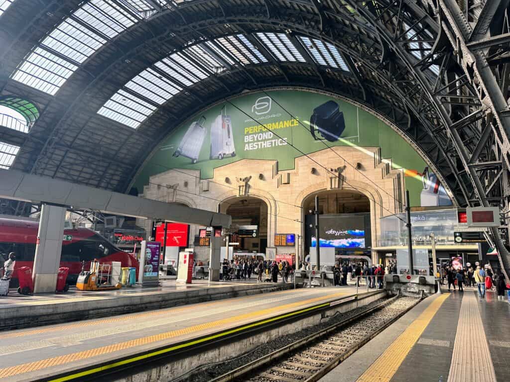 Train and people inside the Milan train station. The ceiling is tall and arched and made of glass and metal.