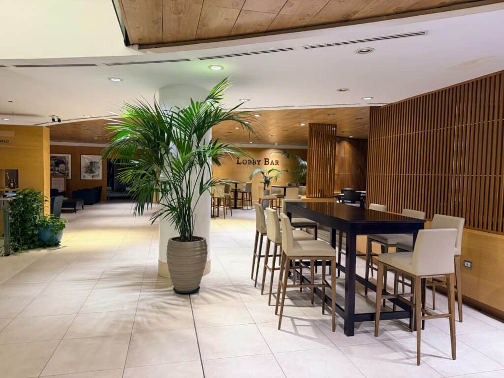 Lobby bar area at Precise House Mantegna Rome. Tall table with bar chairs on right. Tall plant next to it on left. Behind in back are more tables and chairs and sign for 'Lobby Bar.'