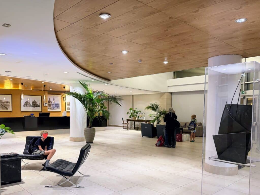 Lobby at Precise House Mantegna Rome. Boy sits on black chair on left. Lobby desk in back left. Woman waits with luggage and child in back right. There are plants in the lobby.