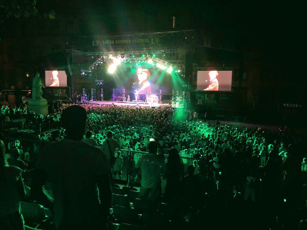 View from seated area of the concert stage of Macklemore performing in Lucca, Italy. A large crowd watches the stage while standing.