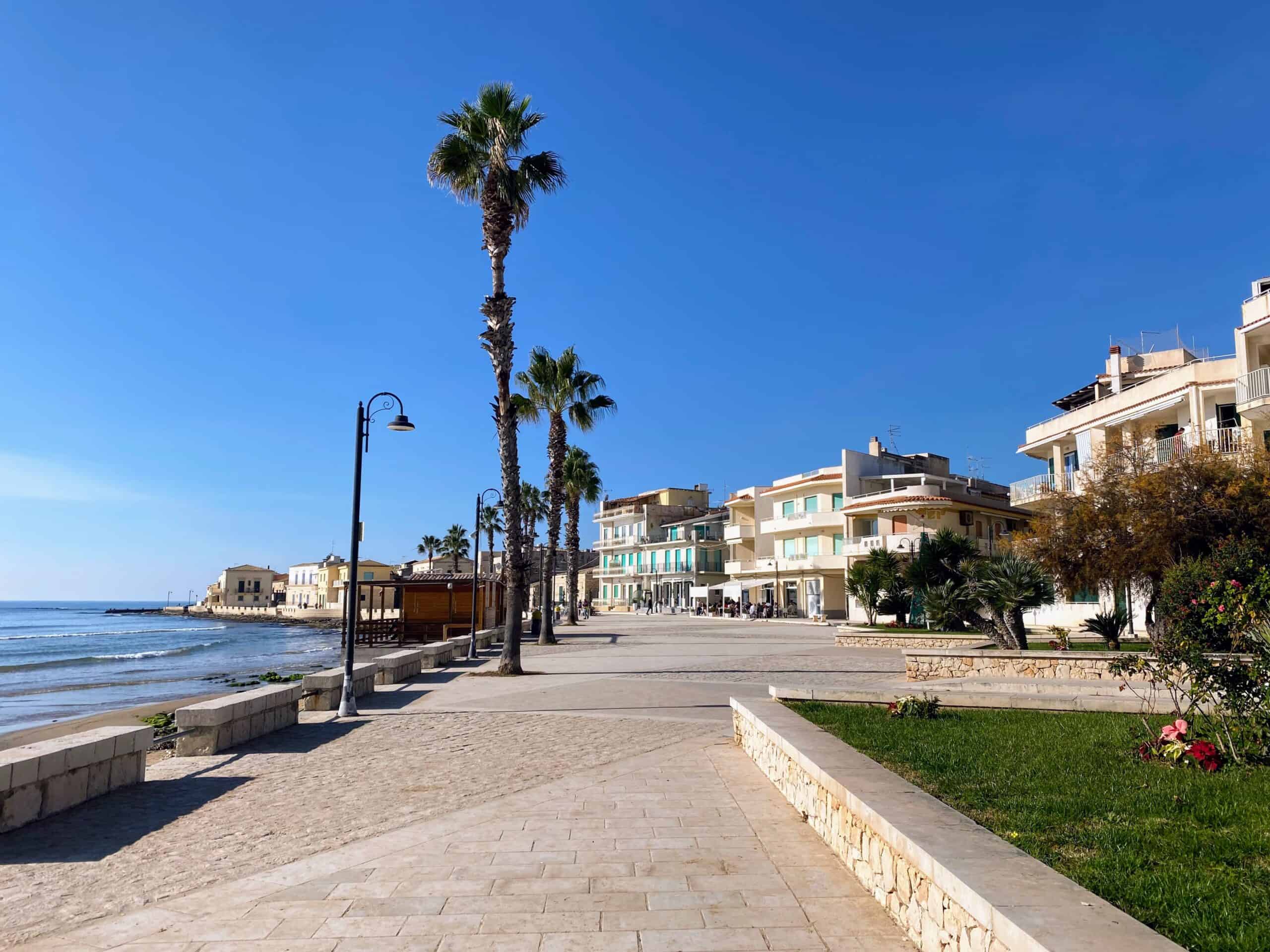 Boardwalk on the sea in Sampieri, Sicily. Palms on the border with the sand, modern buildings on the left side of the boardwalk.
