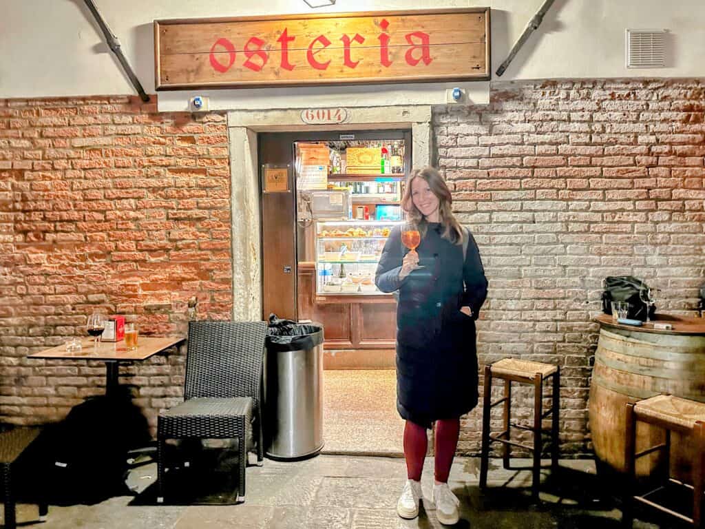 Woman holds up glass of aperol spritz in front of an osteria entrance in Venice, Italy.