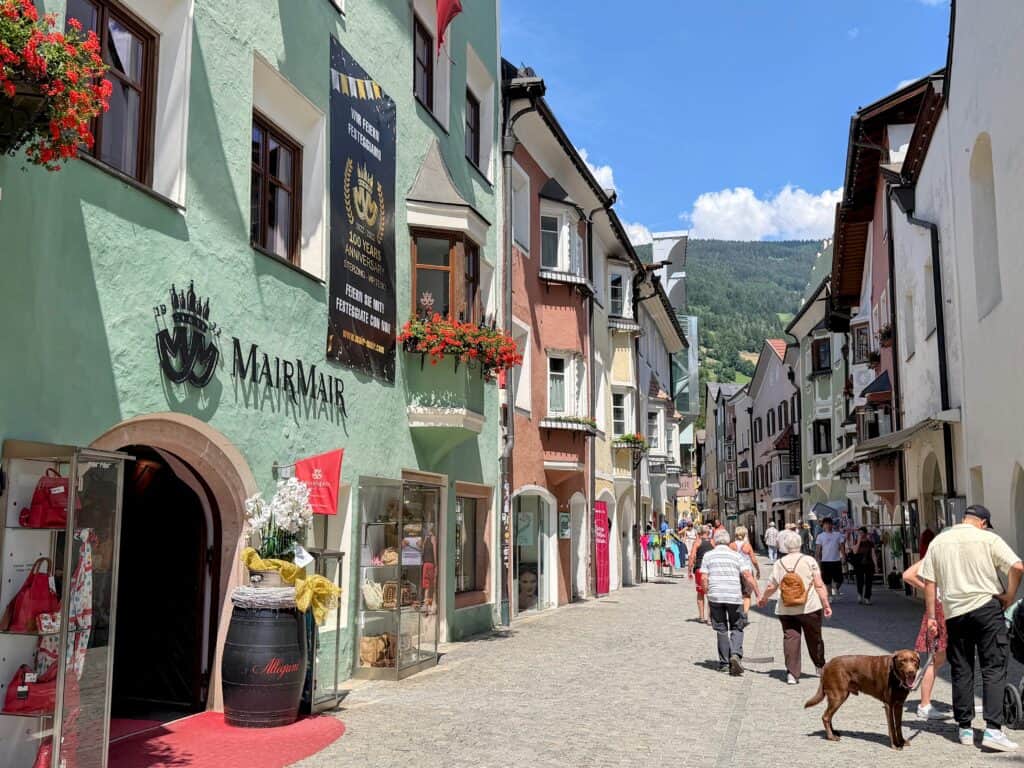 People stand and walk in the pedestrian area of Vipiteno in the Dolomites, Italy. The buildings are painted colorful pastel colors.