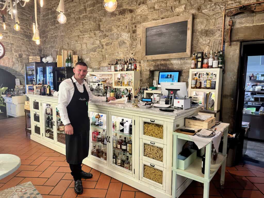 Waiter stands next to white bar with glass windows in Italy. Liquors line the shelves of the bar and waist level and below. He is in a restaurant with stone walls in Italy.