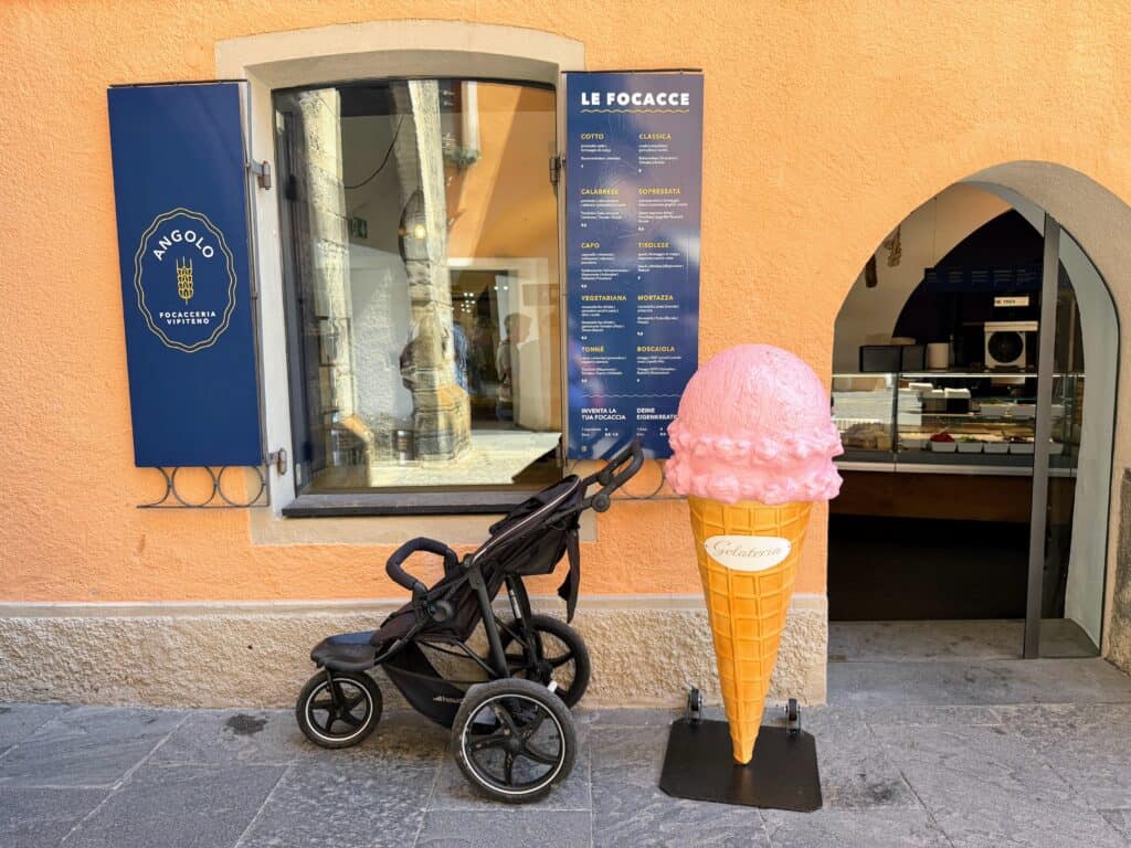A large, plastic gelato cone is in front of the entrance to a sandwich and gelato shop in Vipiteno, Italy. There is a stroller next to it. The menu for the shop is on the wall. You can see a window and the entrance door is open and there are sandwich toppings on display.