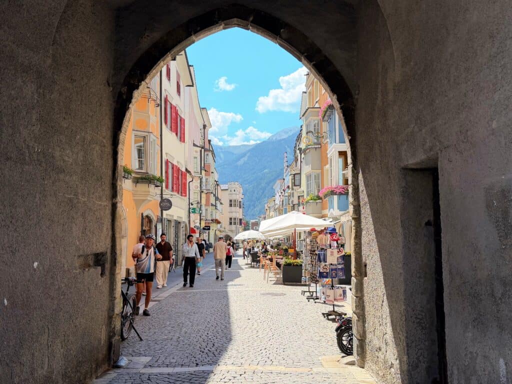An archway at the bottom of the clocktower in Vipiteno, Italy. The view through it is of the cobblestone pedestrian street and the colorful buildings that line each side of it. Sidewalk cafes and people walking on the street.