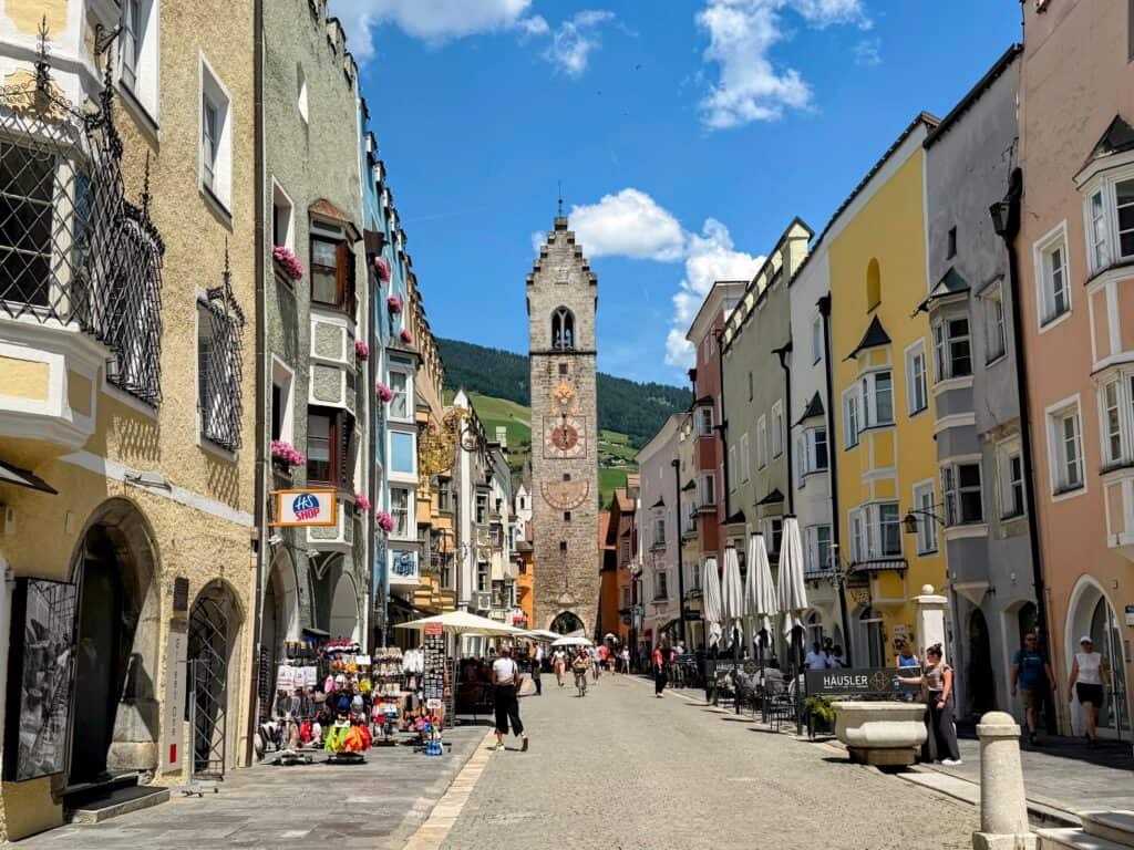Looking at the clocktower of Vipiteno, Italy. Colorful pastel buildings line the cobblestone street that leads up to the clocktower. Green forested hills behind the clock tower. A few people walk on the pedestrian street. There are shops and cafe tables and chairs on the sidewalks. This is in Vipiteno, Italy.