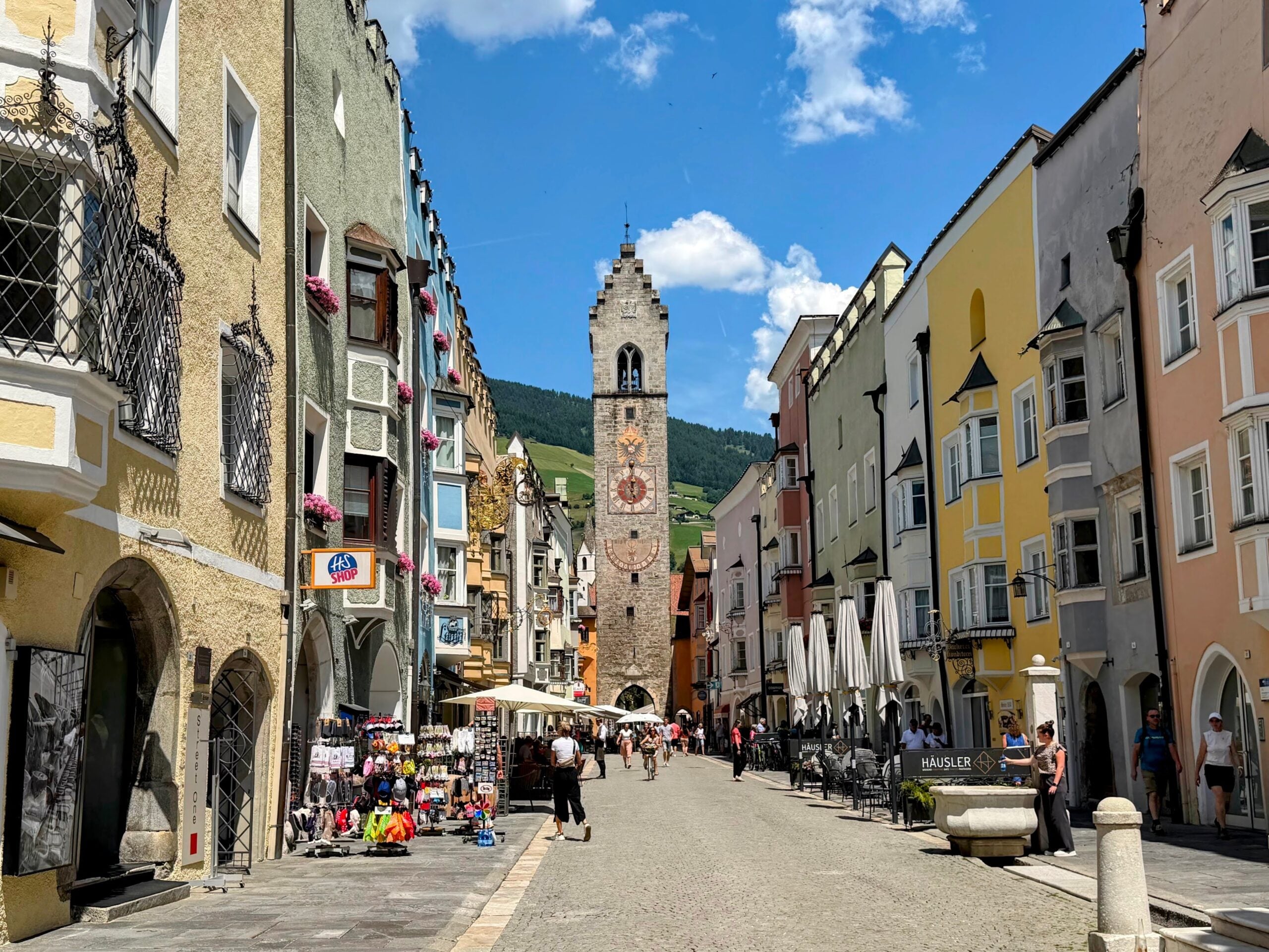 Looking at the clocktower of Vipiteno, Italy. Colorful pastel buildings line the cobblestone street that leads up to the clocktower. Green forested hills behind the clock tower. A few people walk on the pedestrian street. There are shops and cafe tables and chairs on the sidewalks. This is in Vipiteno, Italy.