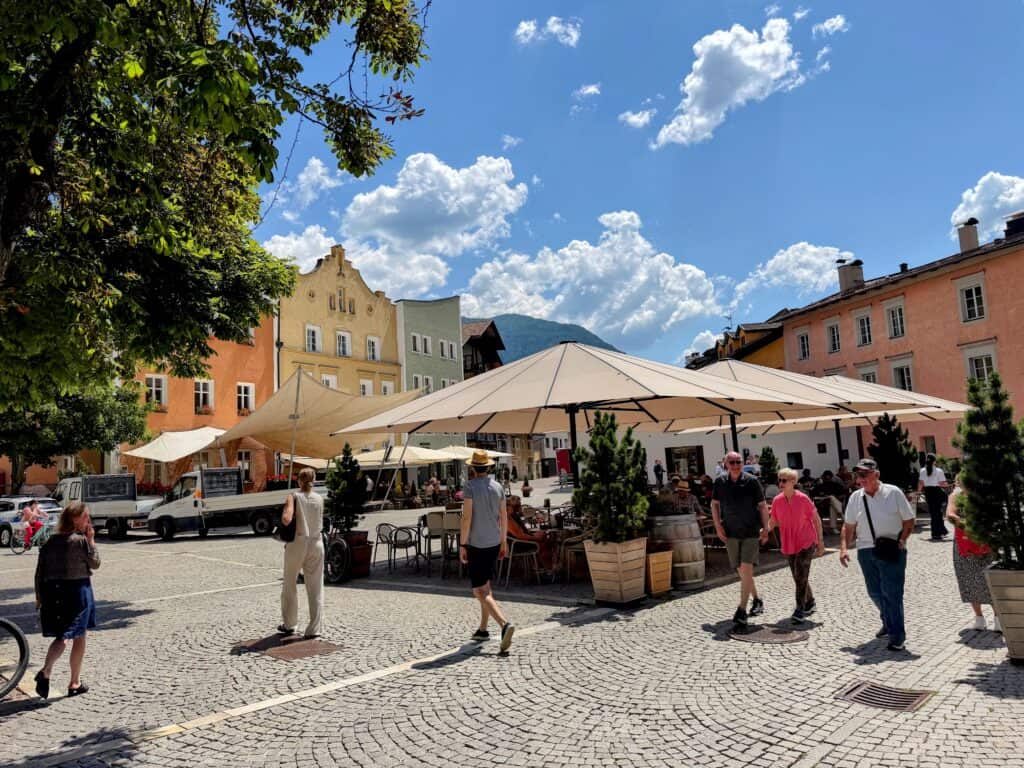 Restaurant in the main piazza of Vipiteno, Italy is covered by large white umbrellas. People walk near the umbrellas and others sit at the tables. The piazza is surrounded by colorful buildings.