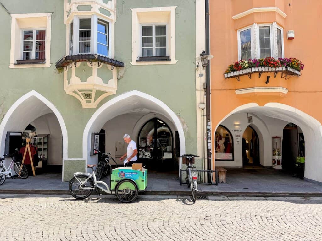 Green and orange building with porticos and shops in the porticoes. Cobblestone street in front. Man looks at his bicycle that has behind seat storage. This is in Vipiteno, Italy.