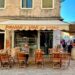 Wooden chairs and tables are set up outside Panini e Vini da Babbo in Venice, Italy. People sit at the tables and others walk by on the wide, pedestrian way. The window displays cicchetti for sale. It's a sunny day with no clouds.